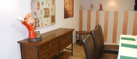A room with a wooden dresser, a chair, and a table with striped tablecloths. There are wall decorations and a lamp.