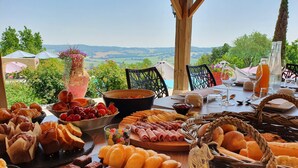 Een ontbijttafel met diverse gebakjes, fruit en drankjes, met uitzicht op een schilderachtig landschap.