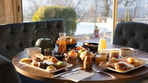 Een ontbijttafel met diverse etenswaren, waaronder gebak, fruit en drankjes, in een kamer met grote ramen en uitzicht op een zwembad.