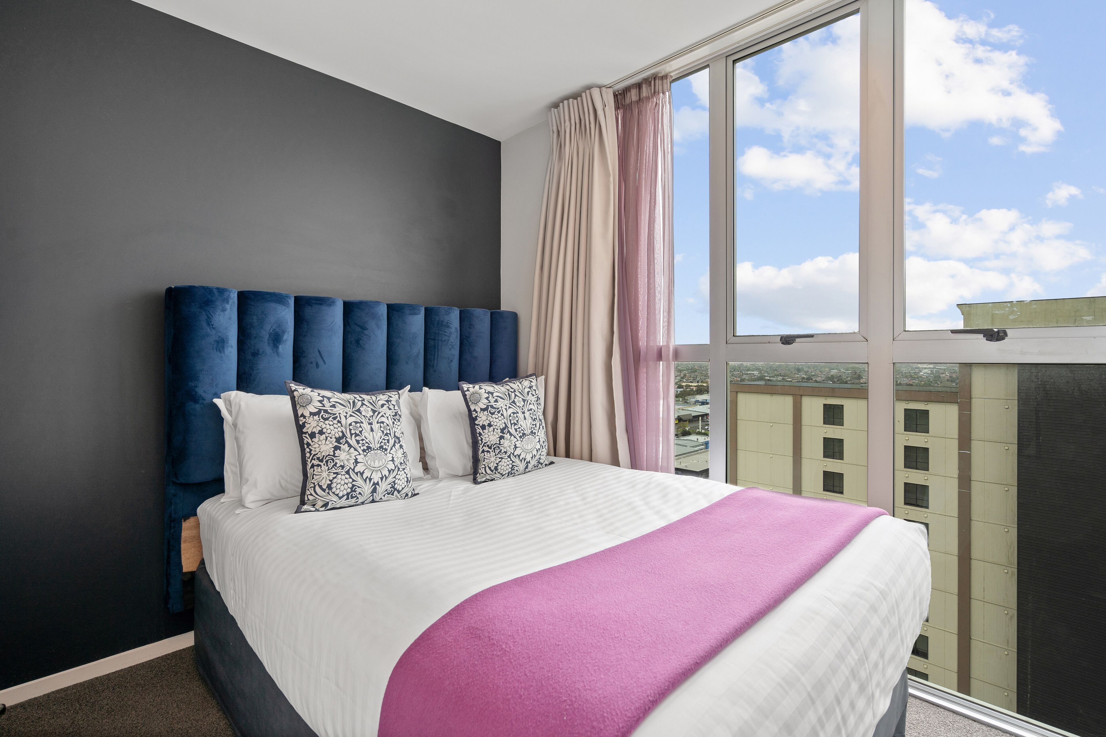 Quarto de hotel moderno com uma cama grande, cabeceira azul escura e vista para a cidade através de janelas amplas.