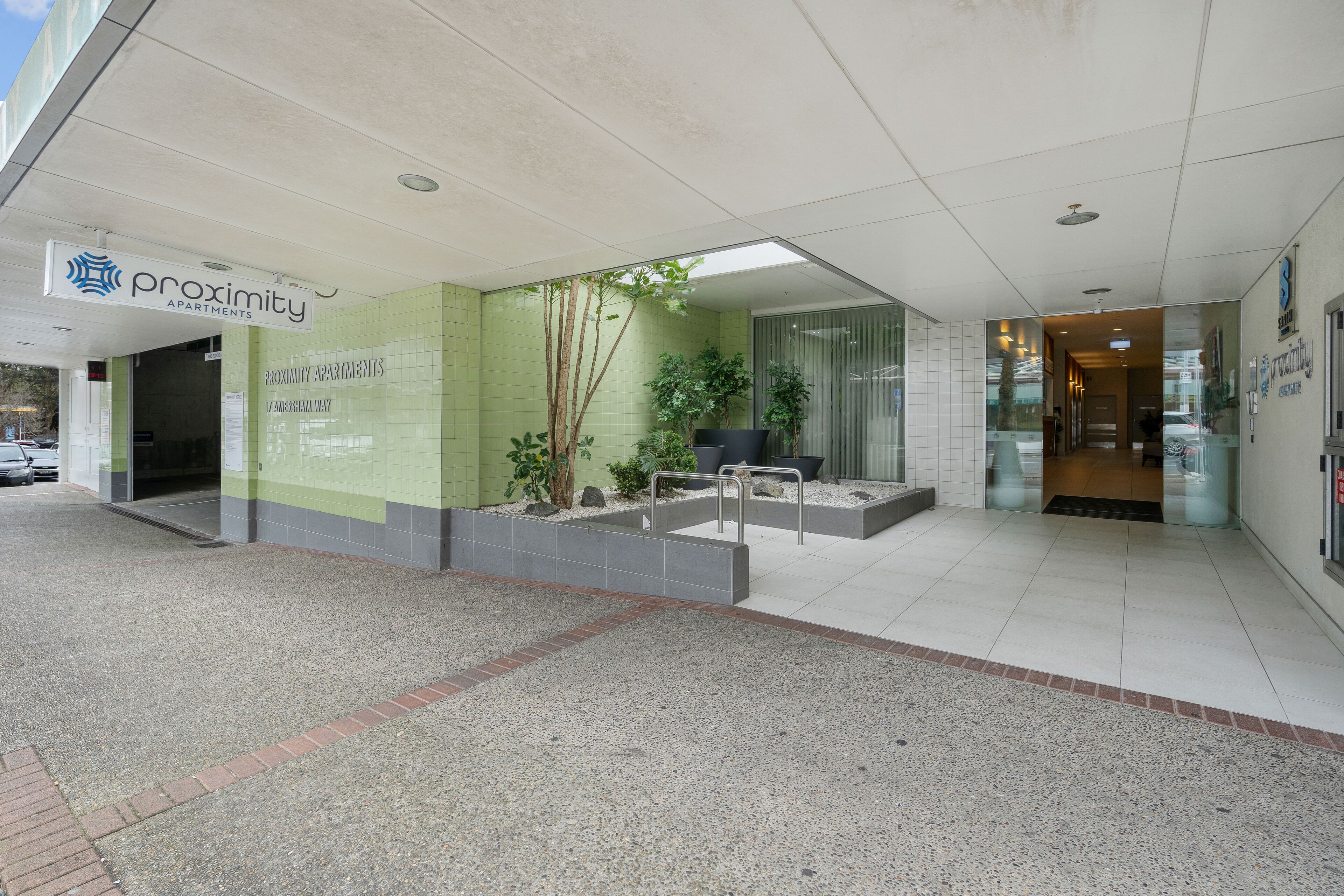 En modern entré till en byggnad med en skylt som visar texten ”Proximity Apartments” och ett välanlagt område med trappor och planterade blommor i krukor.