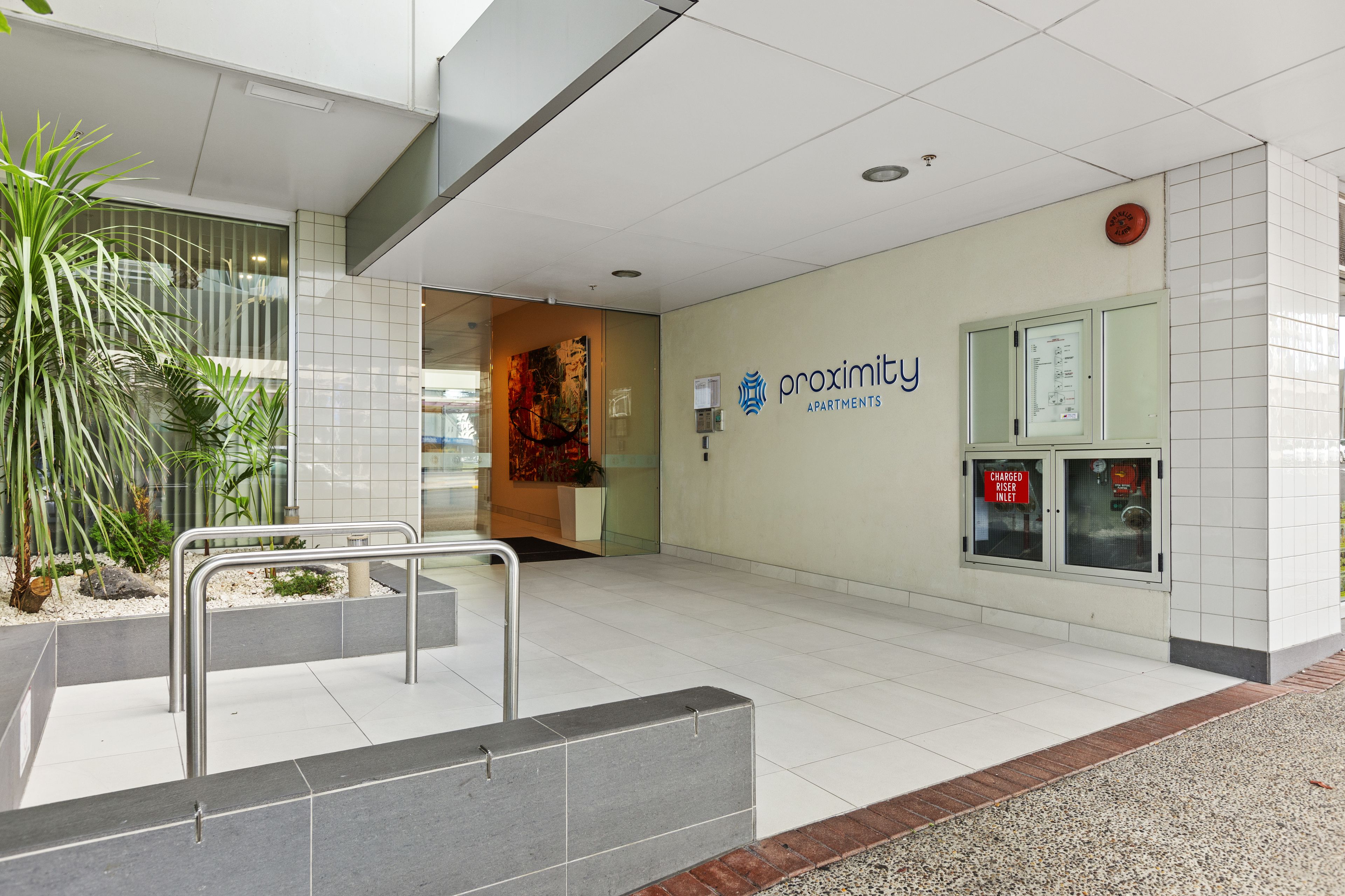 A clean, modern lobby with a reception desk, a sign for 'Proximity Apartments', and a 'Charged Riser Inlet' sign.