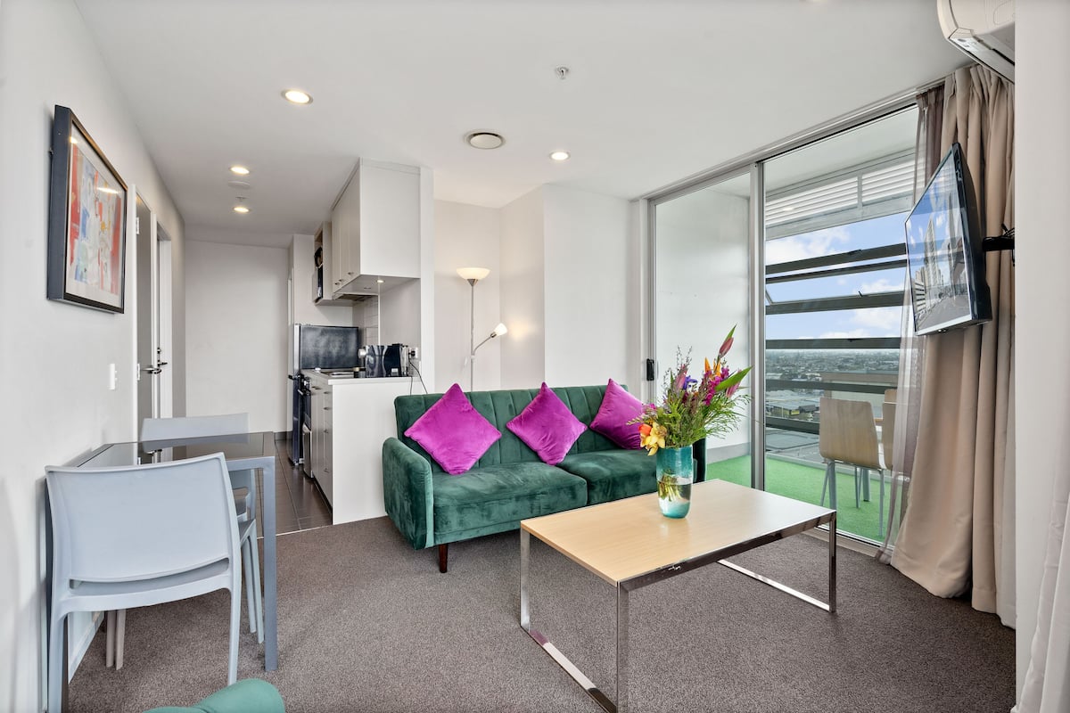 A modern living room with a green sofa, a small table with a vase, and a balcony with a view.