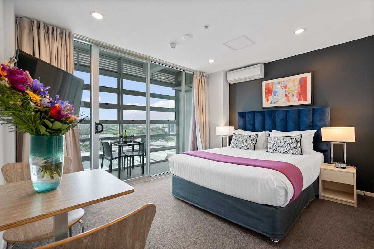 A modern hotel room with a large bed, a desk, and a view of the city through floor-to-ceiling windows.
