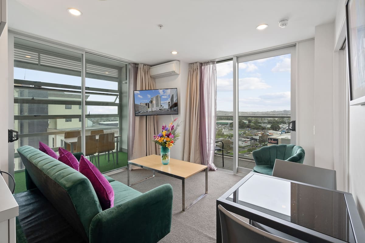 A modern hotel room with a green sofa, a wooden table with a vase of flowers, a flat-screen TV, and large windows offering a city view.