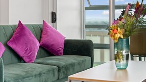 Un canapé vert agrémenté de deux coussins violets, un vase en verre transparent contenant des fleurs et une vue sur le ciel par une fenêtre.