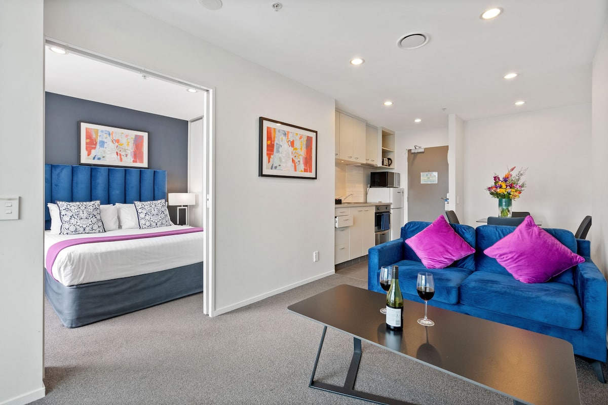 A modern hotel room with a bed, a blue sofa, a coffee table with a wine glass, and a kitchenette in the background.