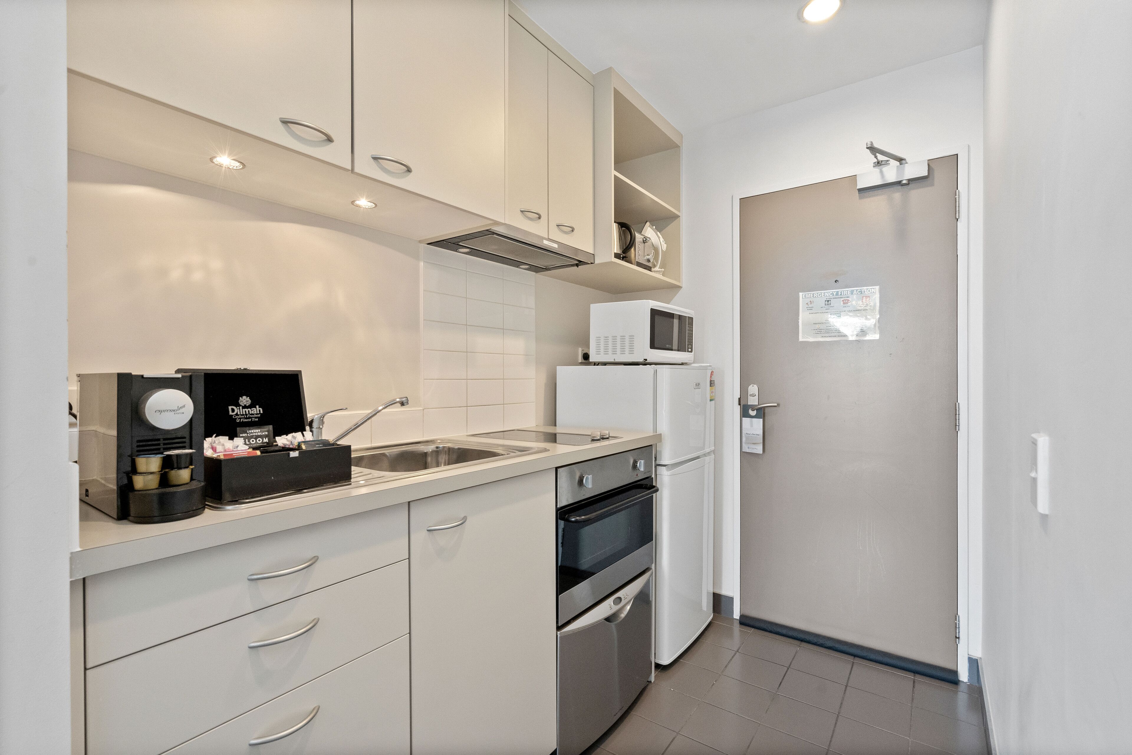 Cocina compacta con armarios blancos, microondas, cafetera y refrigerador.