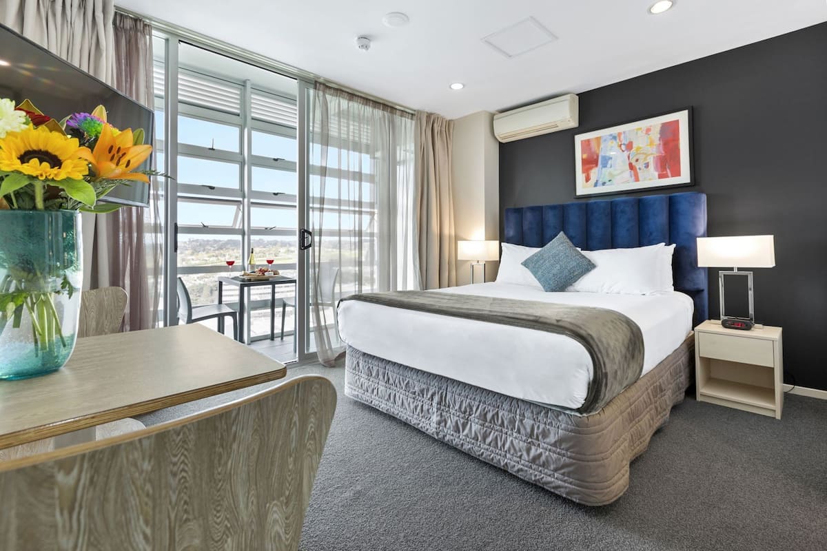 A hotel room with a large bed, a desk, a chair, a vase of flowers, and a view of the outdoors.