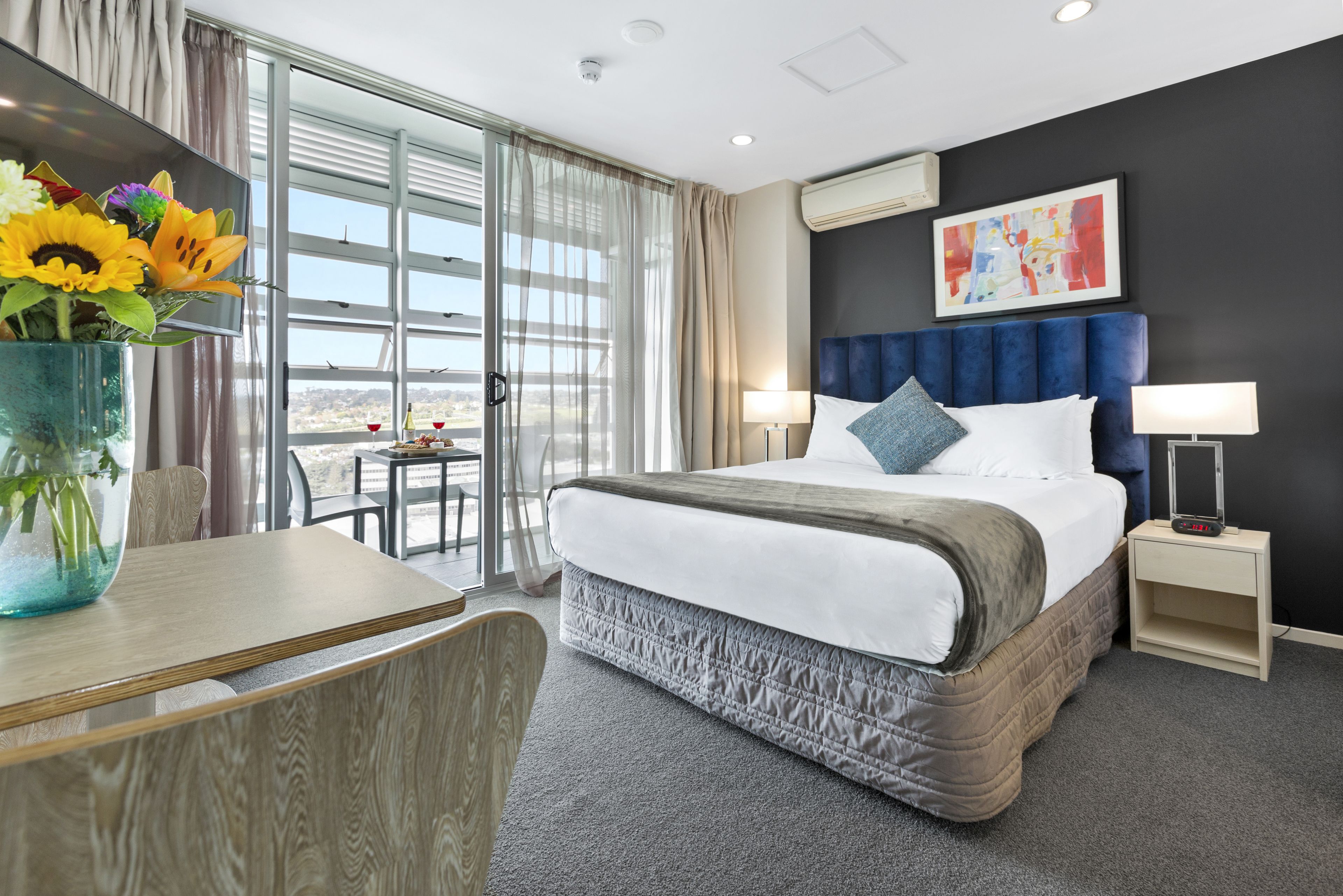A hotel room with a large bed, a desk, a chair, a vase with flowers, a window with curtains, and a painting on the wall.