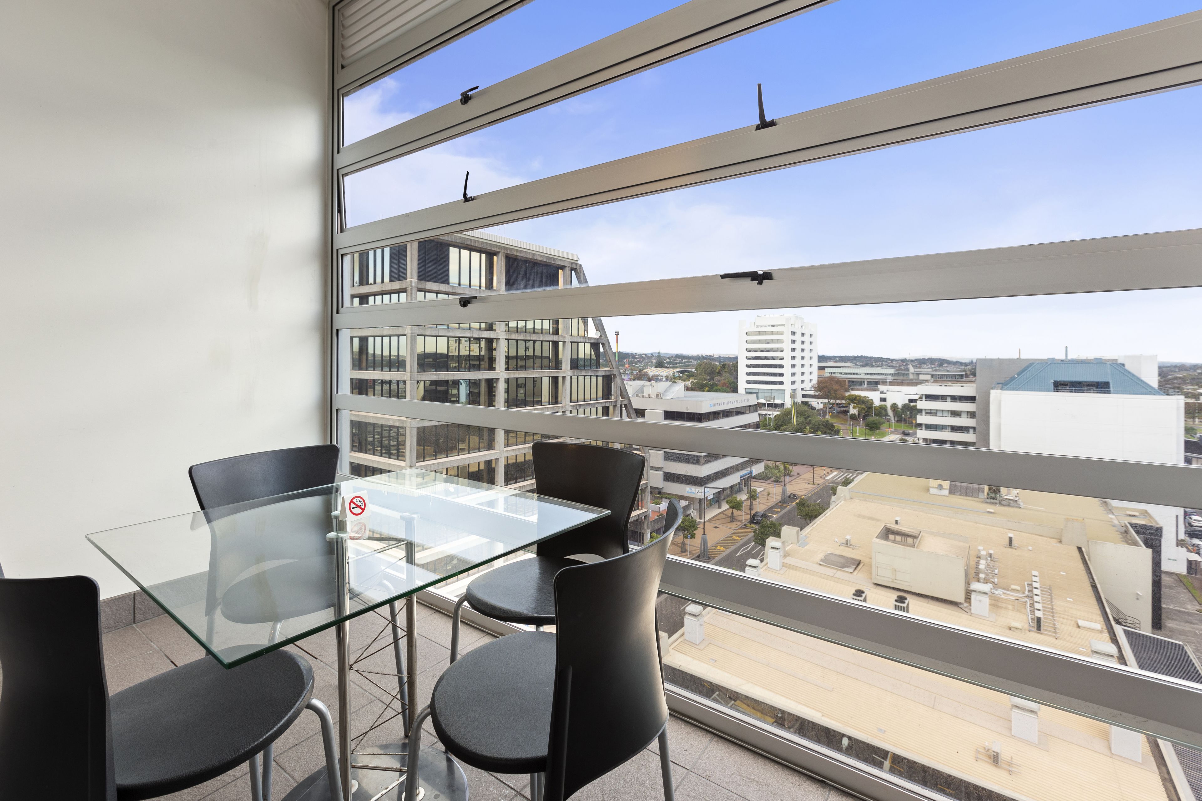 A balcony with a glass table and chairs, overlooking a cityscape with modern buildings.