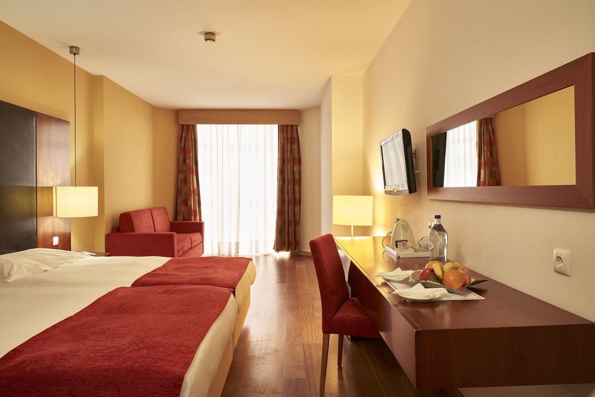 Ein Hotelzimmer mit zwei Betten, einem Schreibtisch, einem Sessel, einem Fernseher und einer Obstschale.