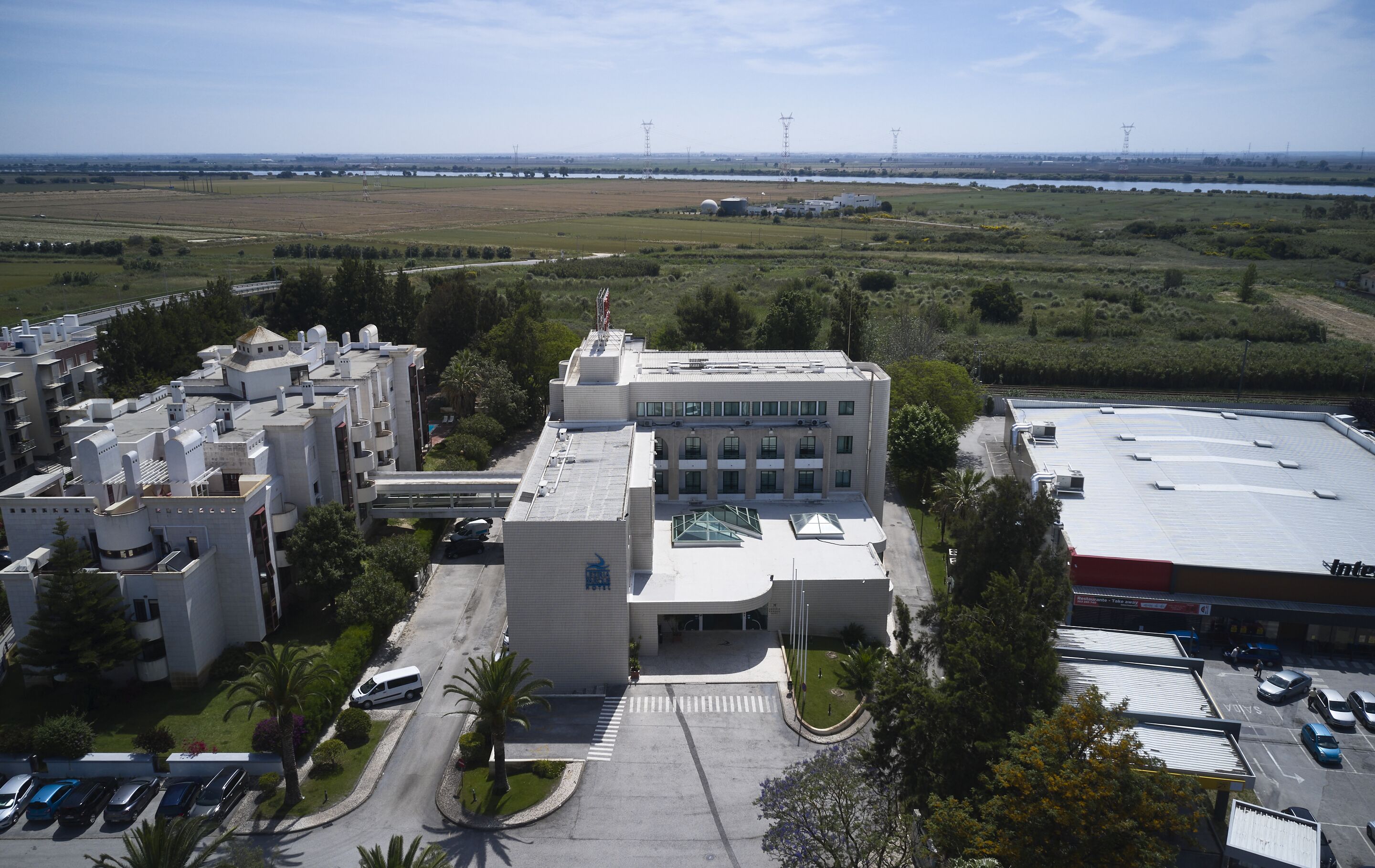 A large building with multiple wings, a parking lot, and a view of a distant landscape including fields and power lines.