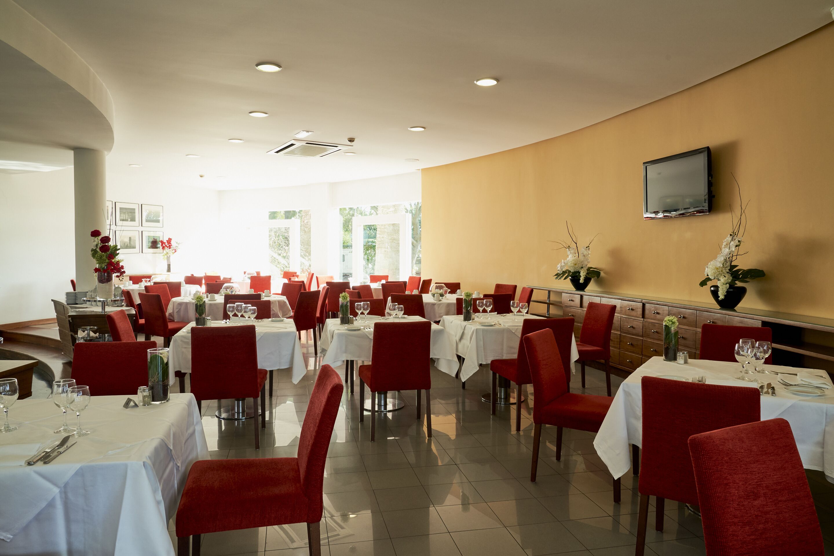 A spacious dining area with multiple tables and chairs, a flat-screen TV mounted on the wall, and large windows allowing natural light.