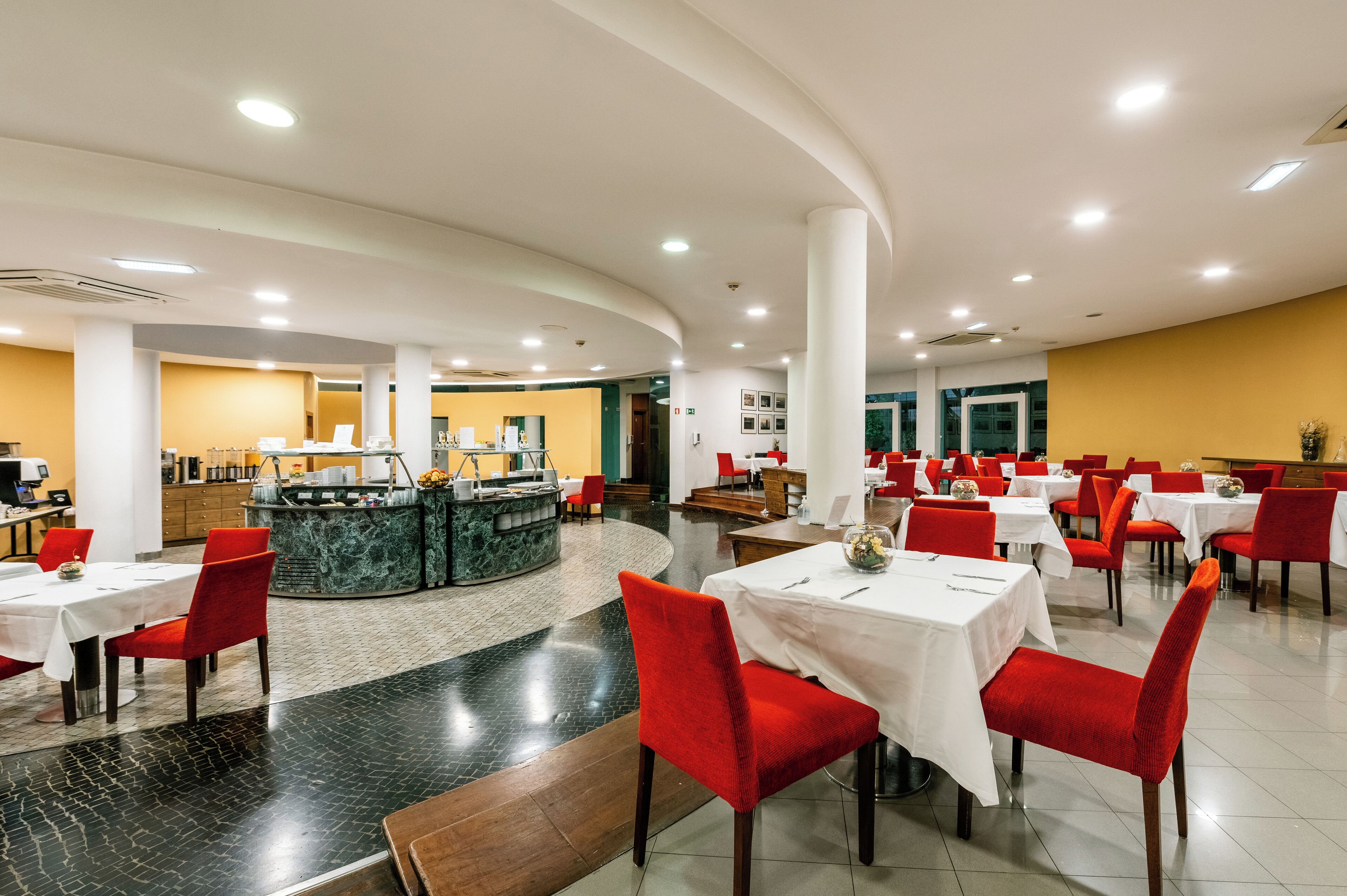 Un vaste espace dédié à la salle à manger, avec de nombreuses tables et des chaises rouges, un comptoir central doté d’un plateau en marbre et un mur d’accent jaune.