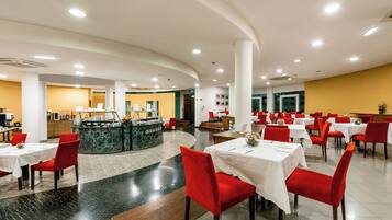 Un vaste espace dédié à la salle à manger, avec de nombreuses tables et des chaises rouges, un comptoir central doté d’un plateau en marbre et un mur d’accent jaune.