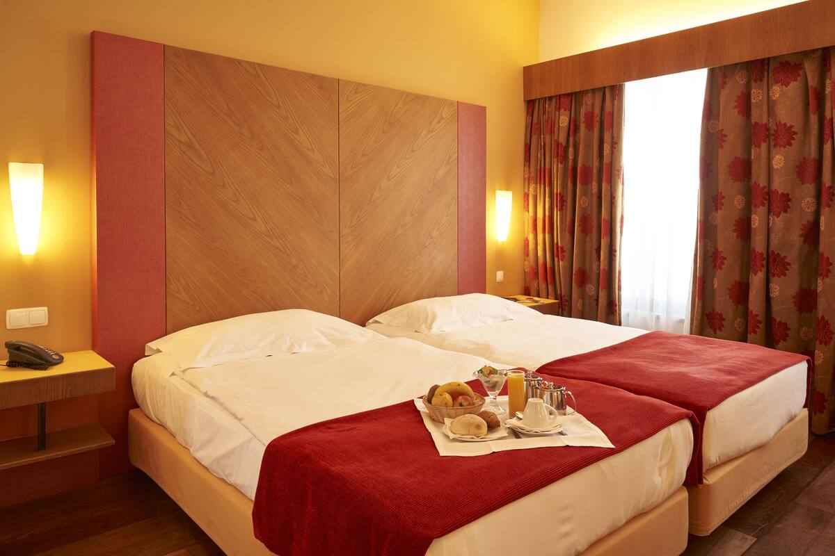 Ein Hotelzimmer mit zwei Betten, einem Nachttisch, einem Telefon und einem Frühstückswagen mit Obst und Getränken.