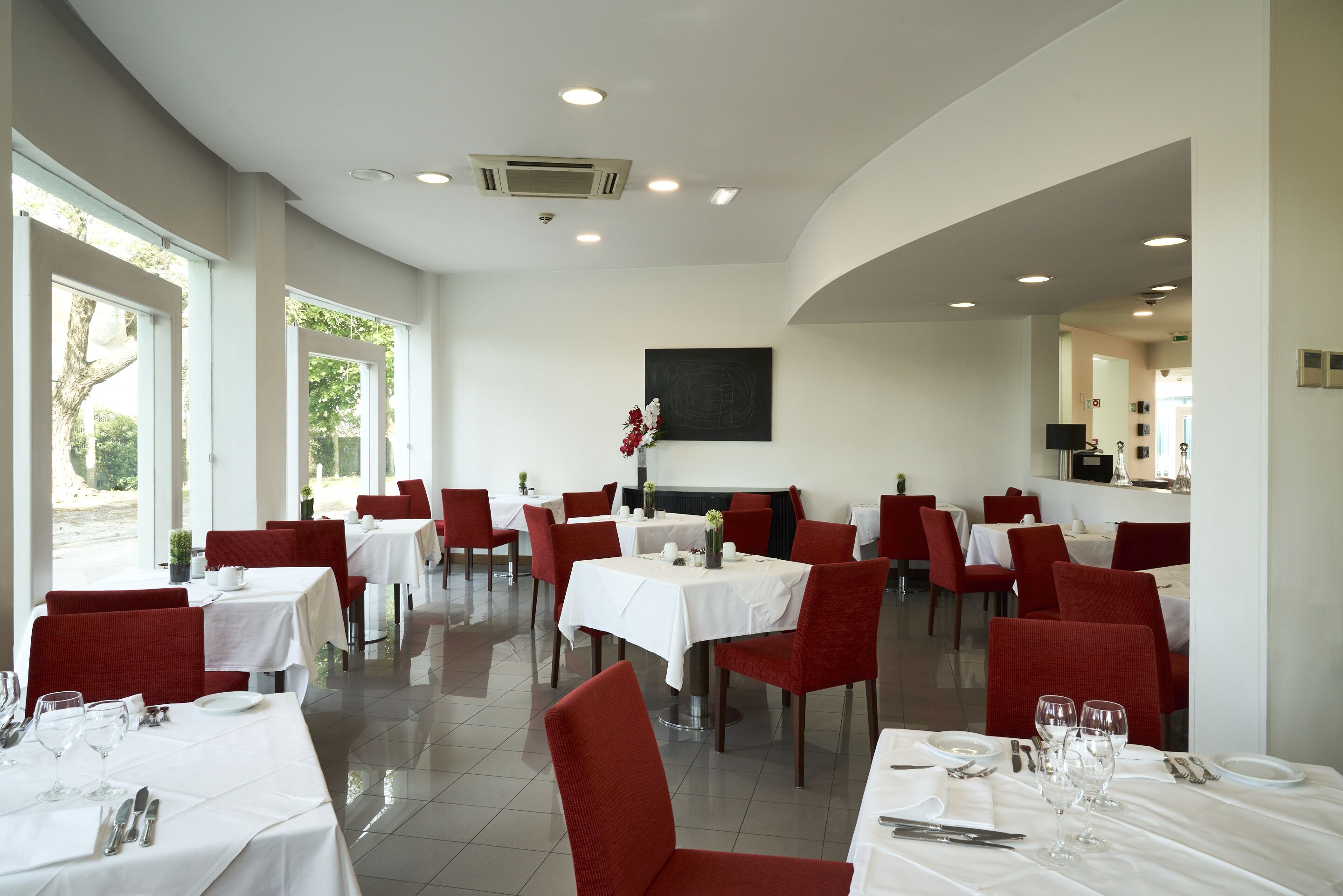 A restaurant with white tables and red chairs, large windows, and a black painting on the wall.