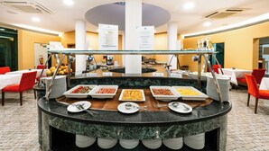 Disposición de bufé con diversos alimentos, una barrera de cristal y mesas dispuestas.