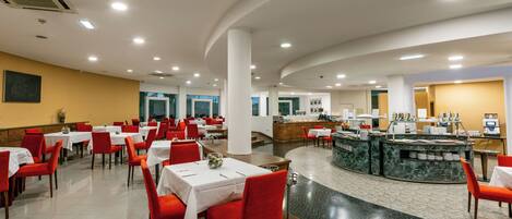 A spacious restaurant with multiple tables and red chairs, a central counter with a marble top, and a tiled floor.