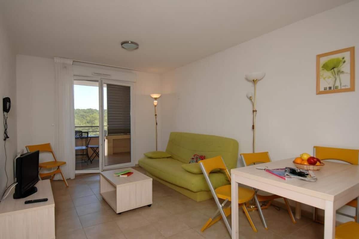 A living room with a green sofa, a white coffee table, a dining table with yellow chairs, a TV, and a balcony with a view.