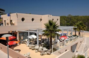 An outdoor seating area with tables and chairs, a palm tree, and a building with circular windows. - Les Parasols D'argens (Roquebrune-sur-Argens)