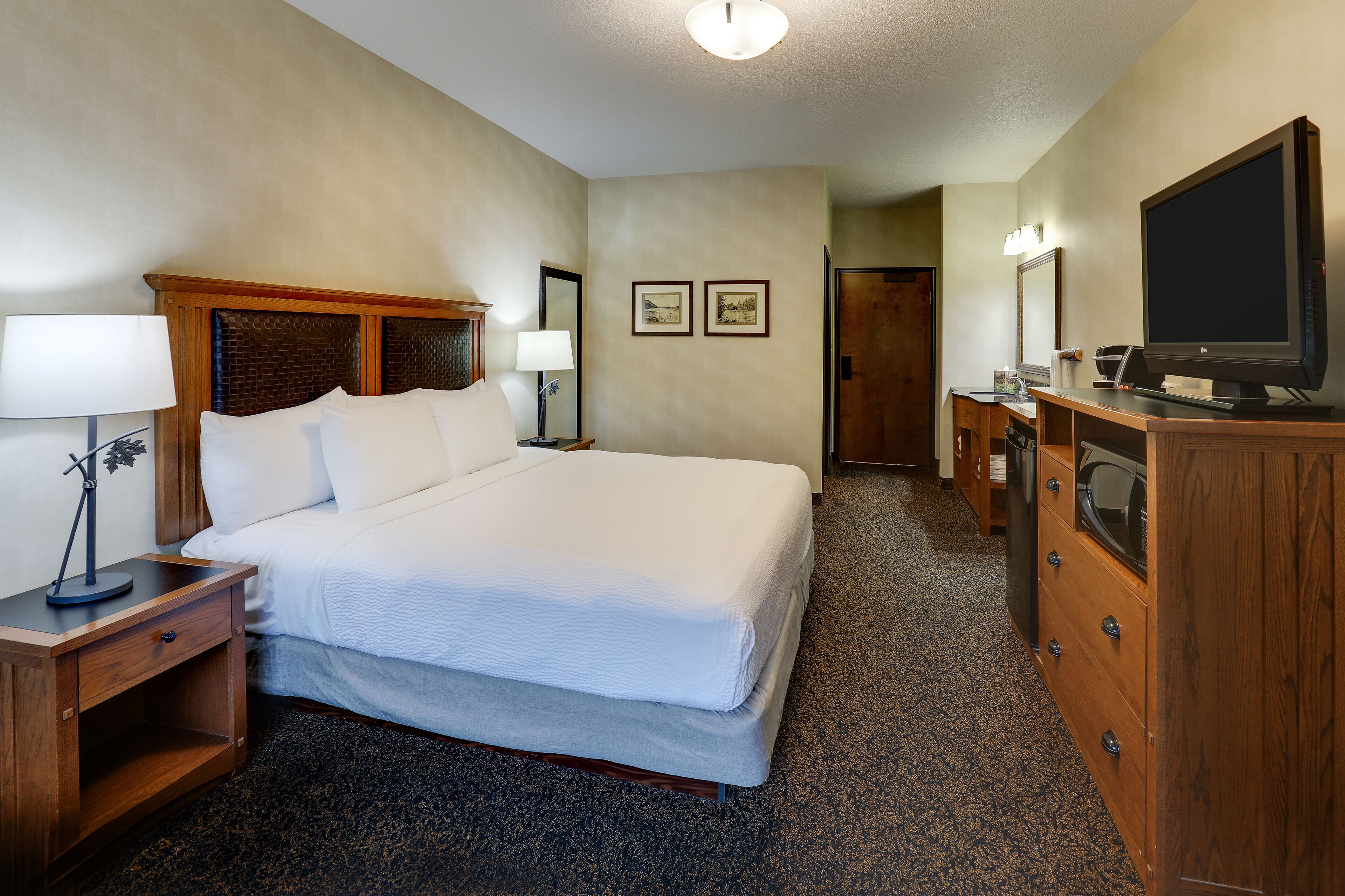 A hotel room with a large bed, wooden headboard, bedside tables with lamps, a dresser with a flat-screen TV, and framed pictures on the wall.