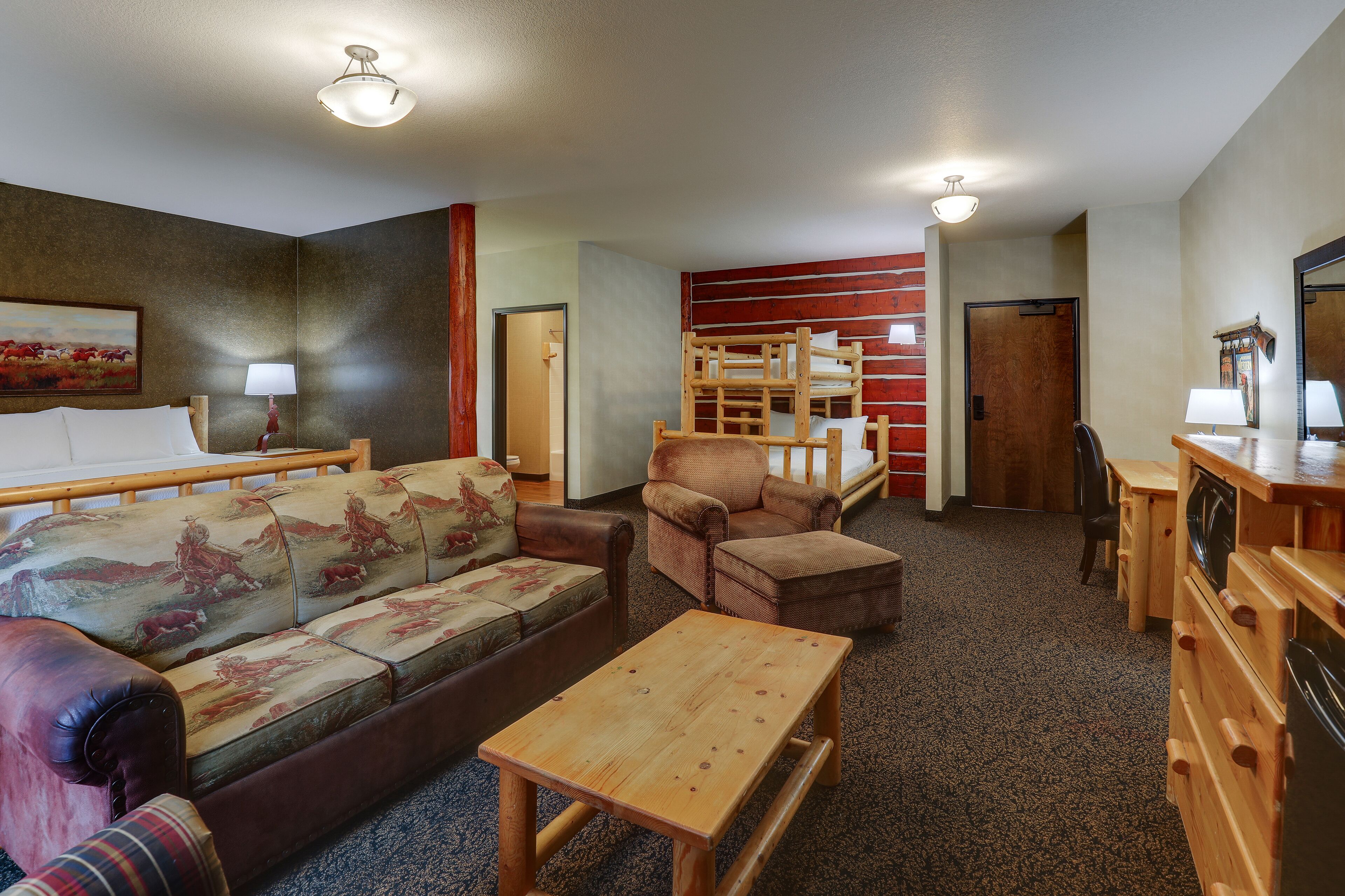 A hotel room with a sofa, armchair, wooden table, and a small wooden desk with a mirror and lamp.