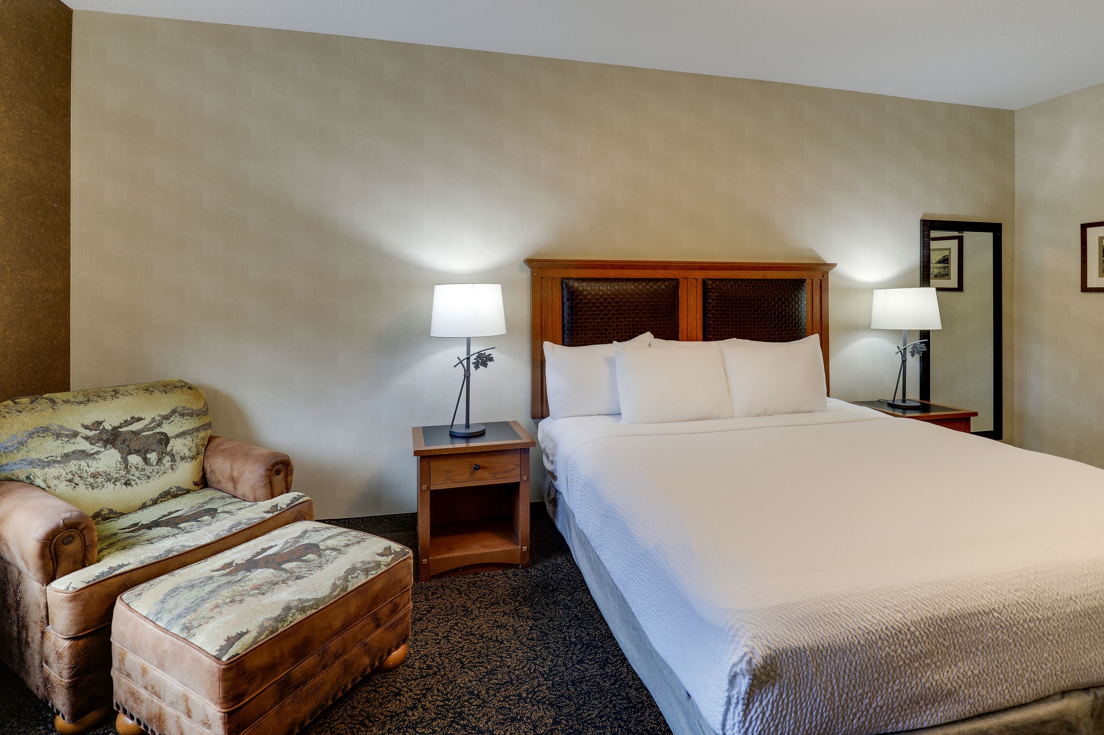 A hotel room with a large bed, a wooden nightstand with a lamp, a chair with a patterned cushion, and a small wooden chest.