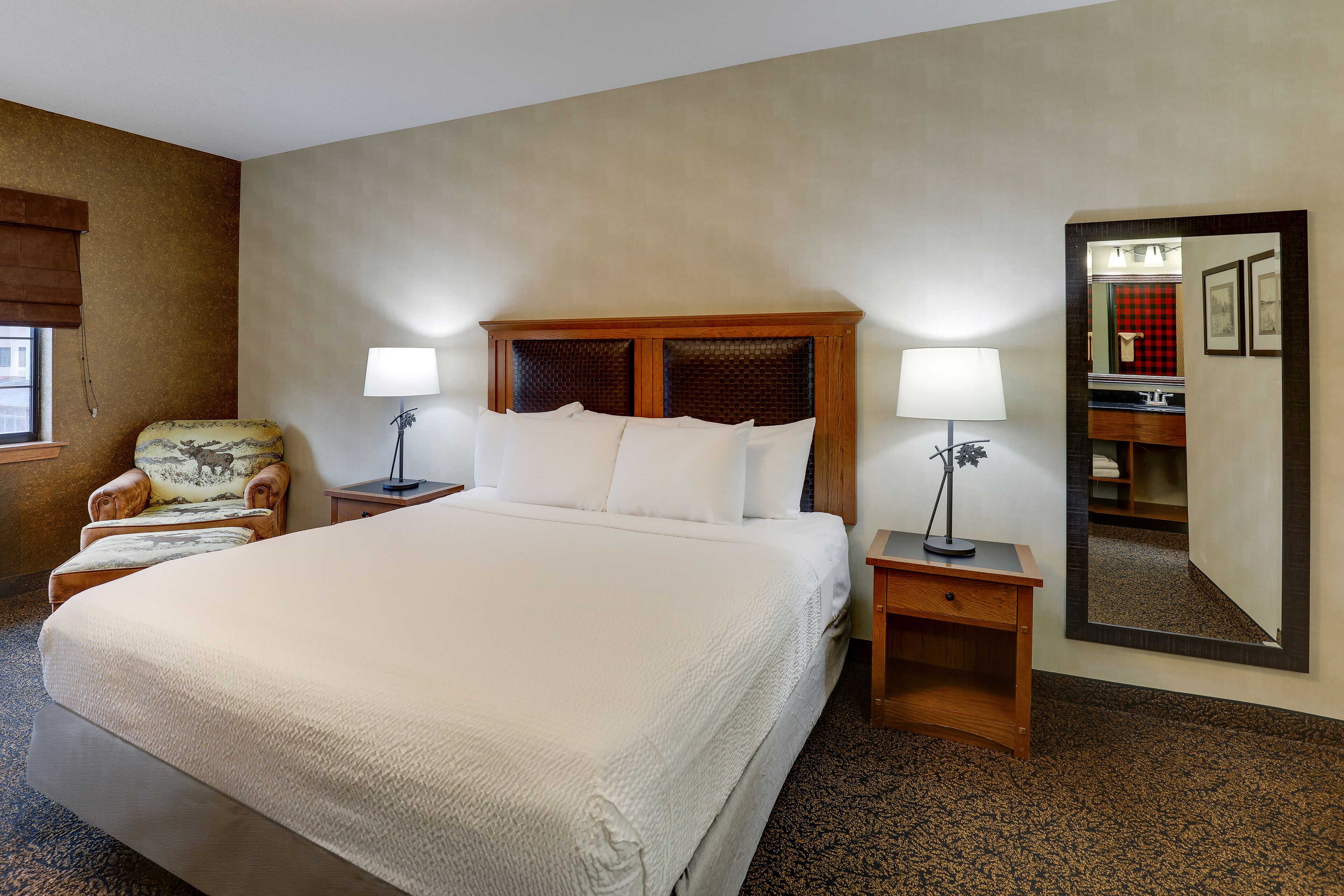 Un cuarto de hotel con una cama grande, dos mesitas de noche con lámparas, una silla con cojín, una mesita de madera y un espejo grande en la pared.
