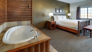 A hotel room with a large bathtub, a wooden bed frame, two bedside tables with lamps, and a view of the city through a window. - Stoney Creek Hotel Sioux City (Sioux City)