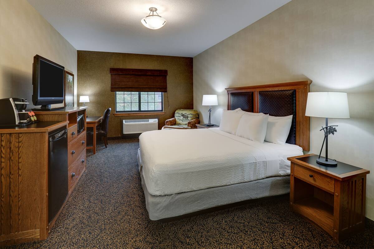 Une chambre d’hôtel comprenant un lit, un bureau avec une télévision, une chaise, une lampe et une fenêtre avec des stores.