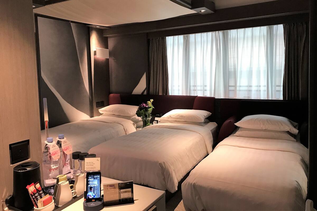 Une chambre d’hôtel avec deux lits, une table de chevet équipée de divers accessoires, une grande fenêtre avec des rideaux et un design moderne au niveau du plafond.