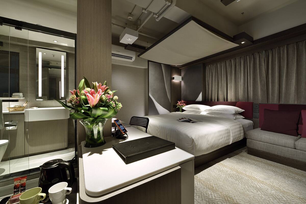 Une chambre d’hôtel moderne, équipée d’un lit, d’une table de chevet et d’un bureau agrémenté d’un vase de fleurs.
