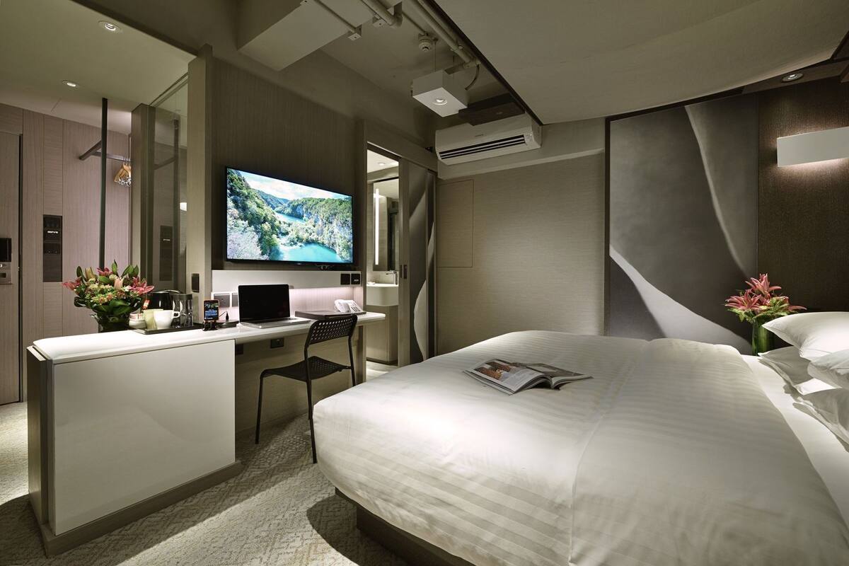 Une chambre d’hôtel avec un lit, un bureau avec un ordinateur portable, une télévision et un vase rempli de fleurs.