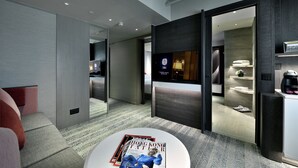 Una habitación de hotel moderna con sofá, una mesa redonda con revistas, televisor y minibar.