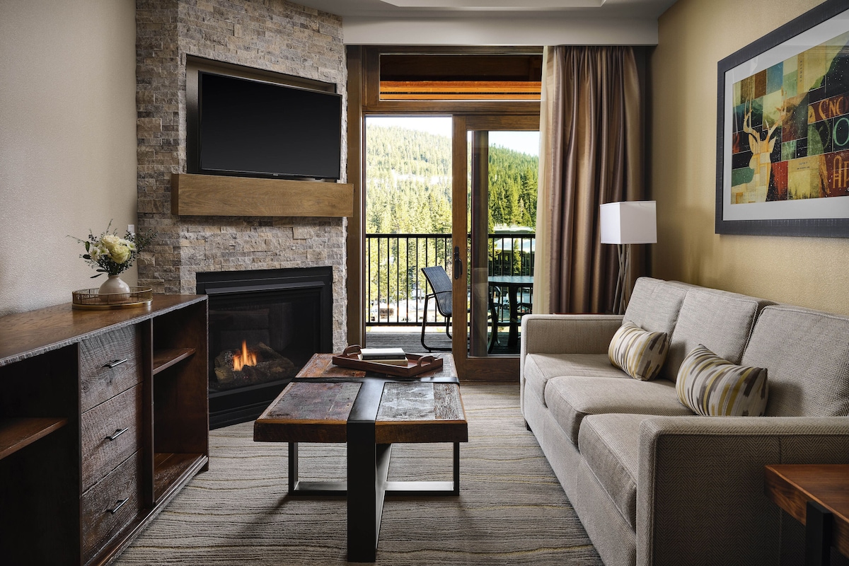 A modern living room with a fireplace, a sofa, a coffee table, and a balcony with outdoor seating.