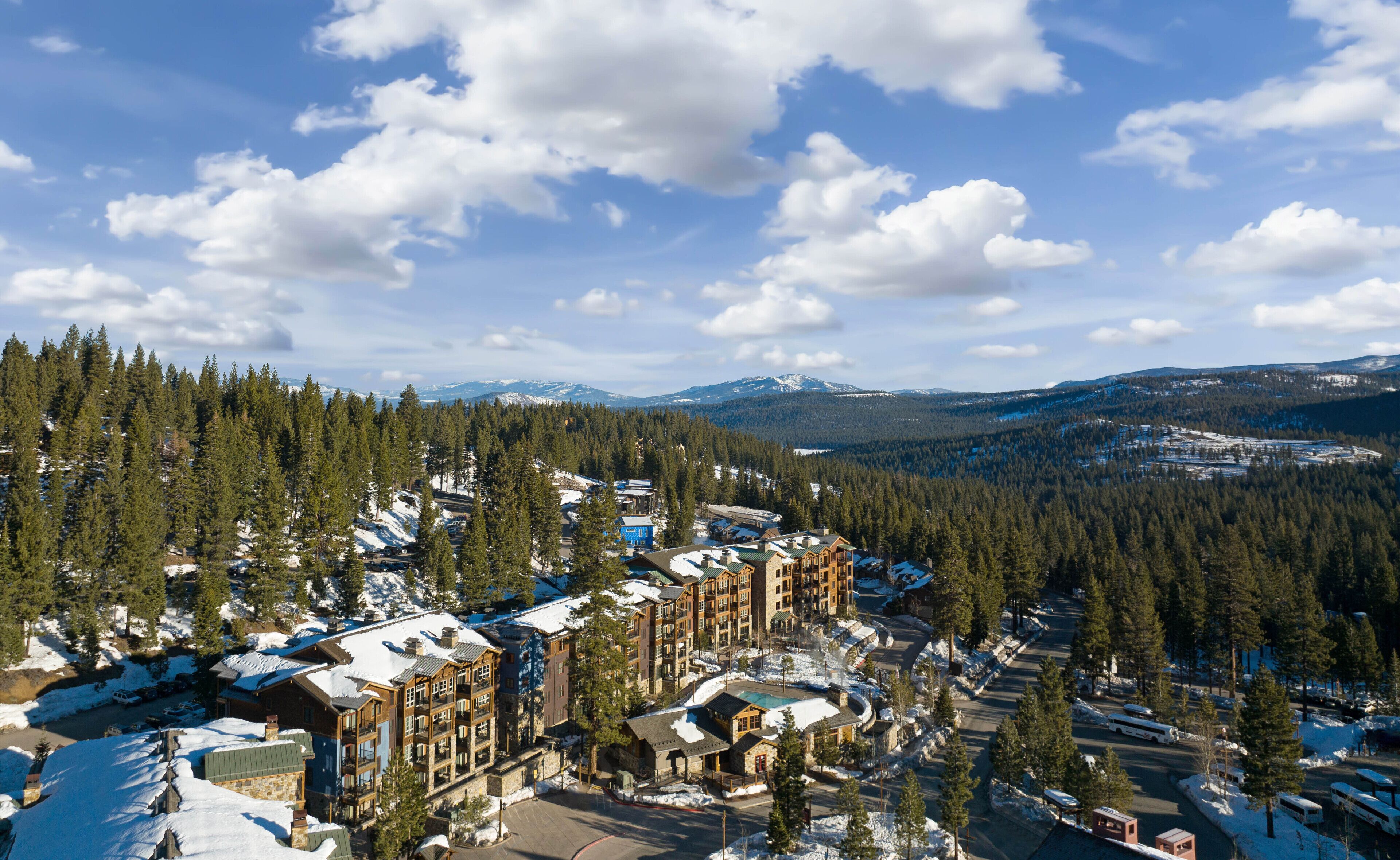 A snow-covered lodge with multiple buildings, surrounded by dense forests and mountains in the background.