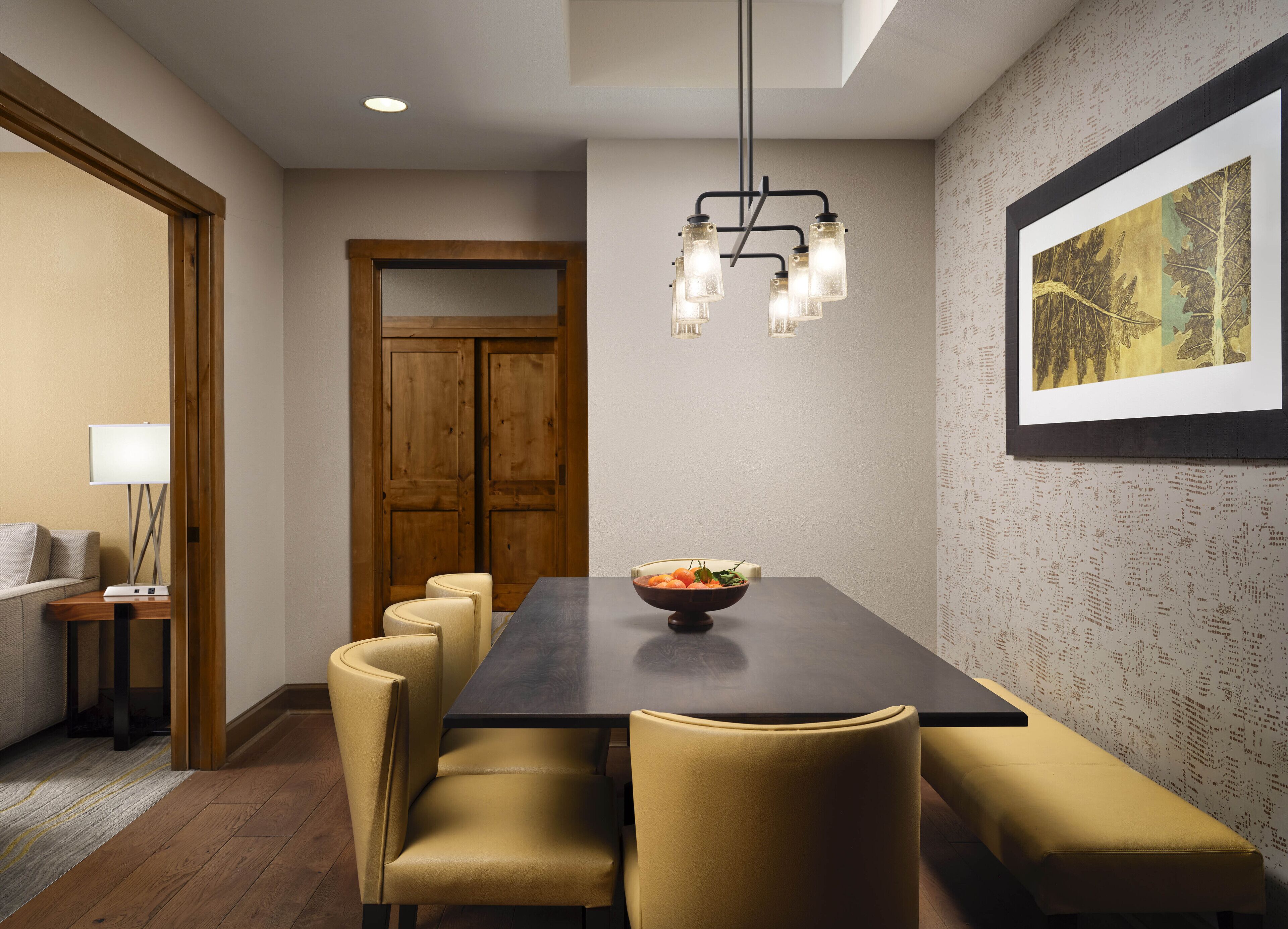 Un comedor con una mesa de madera oscura, sillas tapizadas en amarillo, un frutero y una lámpara colgante moderna.