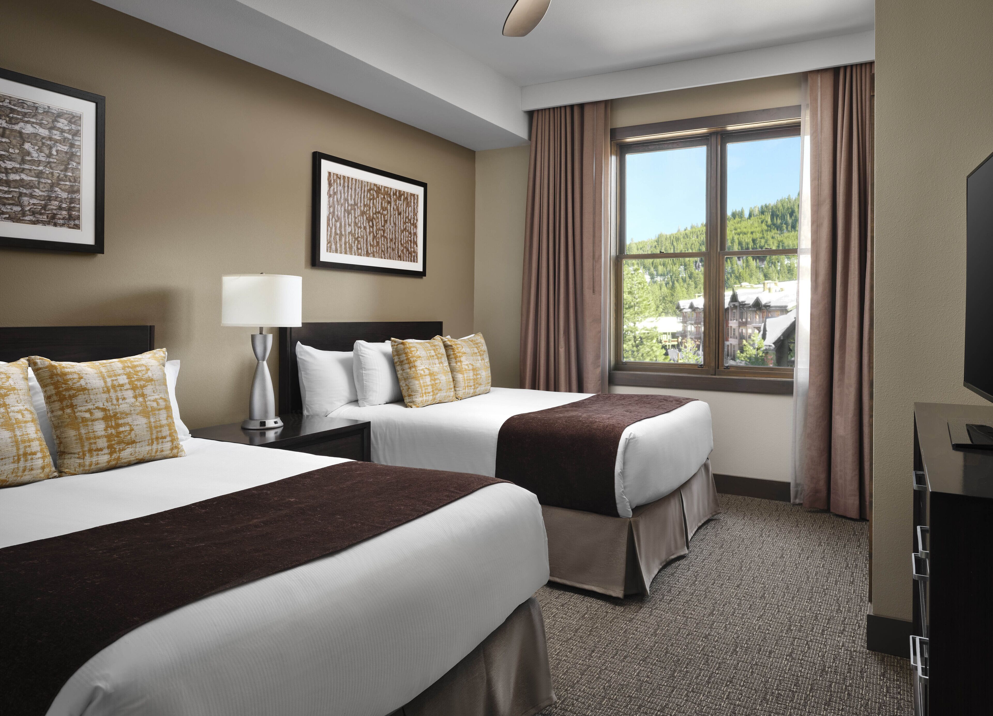 Une chambre d’hôtel avec deux lits, une grande fenêtre avec des rideaux, un téléviseur à écran plat et des œuvres d’art accrochées aux murs.