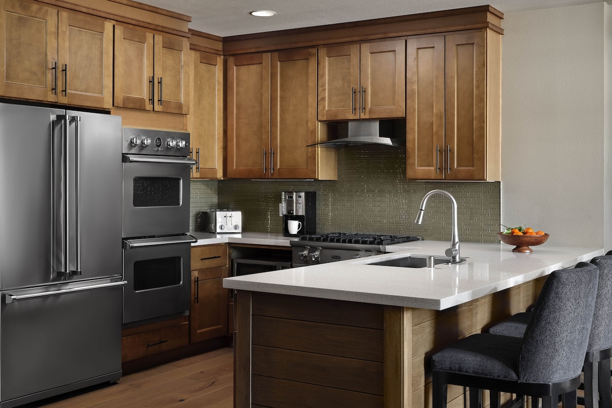 A modern kitchen with wooden cabinets, stainless steel appliances, a central island with bar stools, and a tiled backsplash.