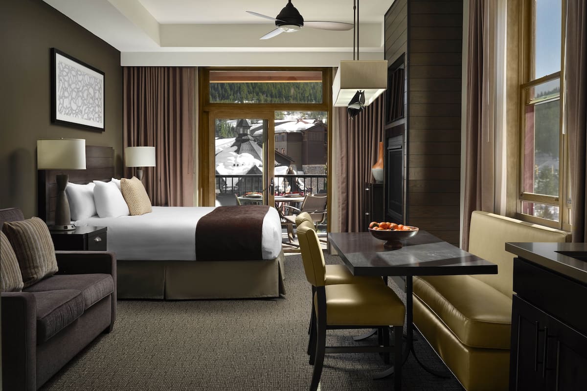 A hotel room with a bed, a desk with a bowl of fruit, a sofa, a chair, a lamp, and a view of the outdoors.