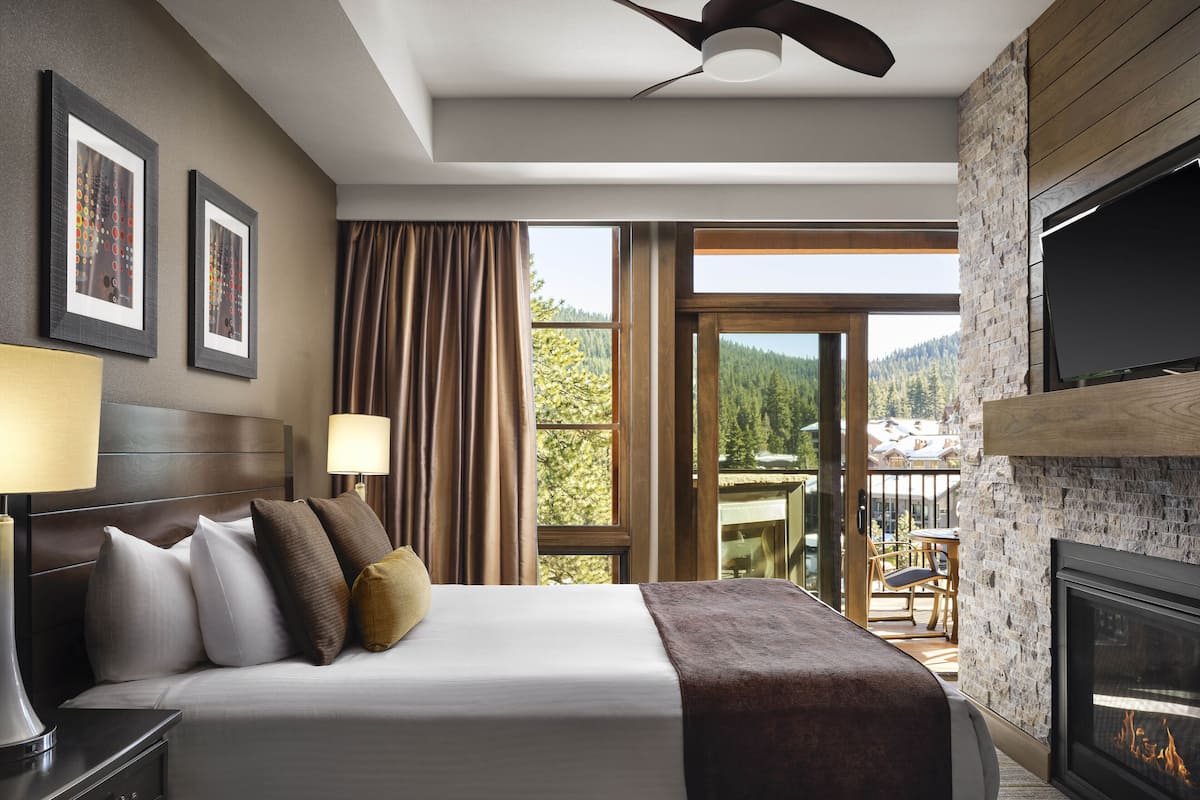A hotel room with a bed, a TV, a fireplace, and a balcony with a view of the mountains.