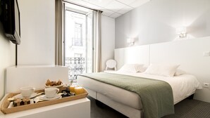 Une chambre d’hôtel avec un lit, une table de chevet avec des articles pour le petit-déjeuner, une fenêtre donnant sur un immeuble et un téléviseur fixé au mur.