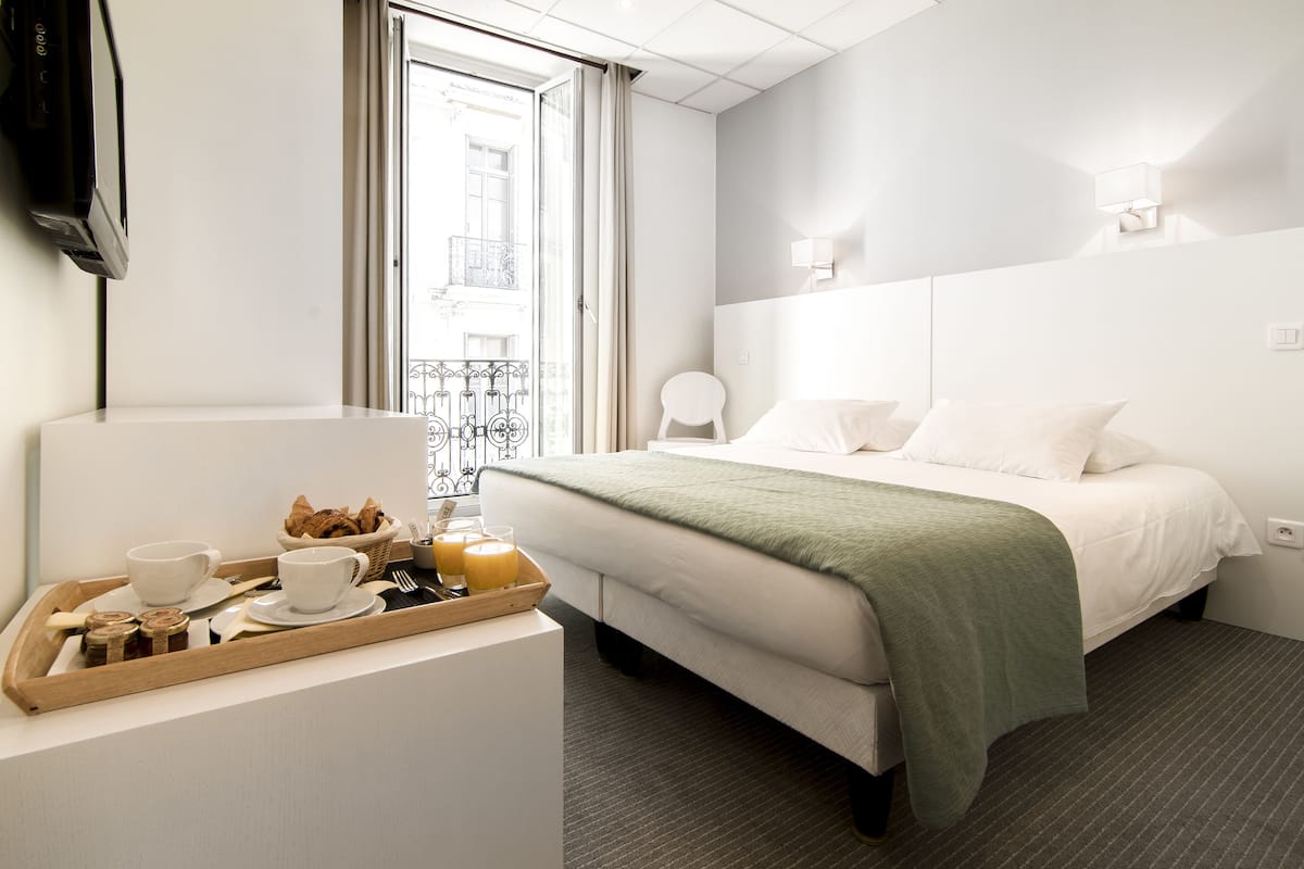 Ein Hotelzimmer mit Bett, Nachttisch mit Frühstücksgebäck, Fenster mit Blick auf ein Gebäude und einem an der Wand befestigten Fernseher.