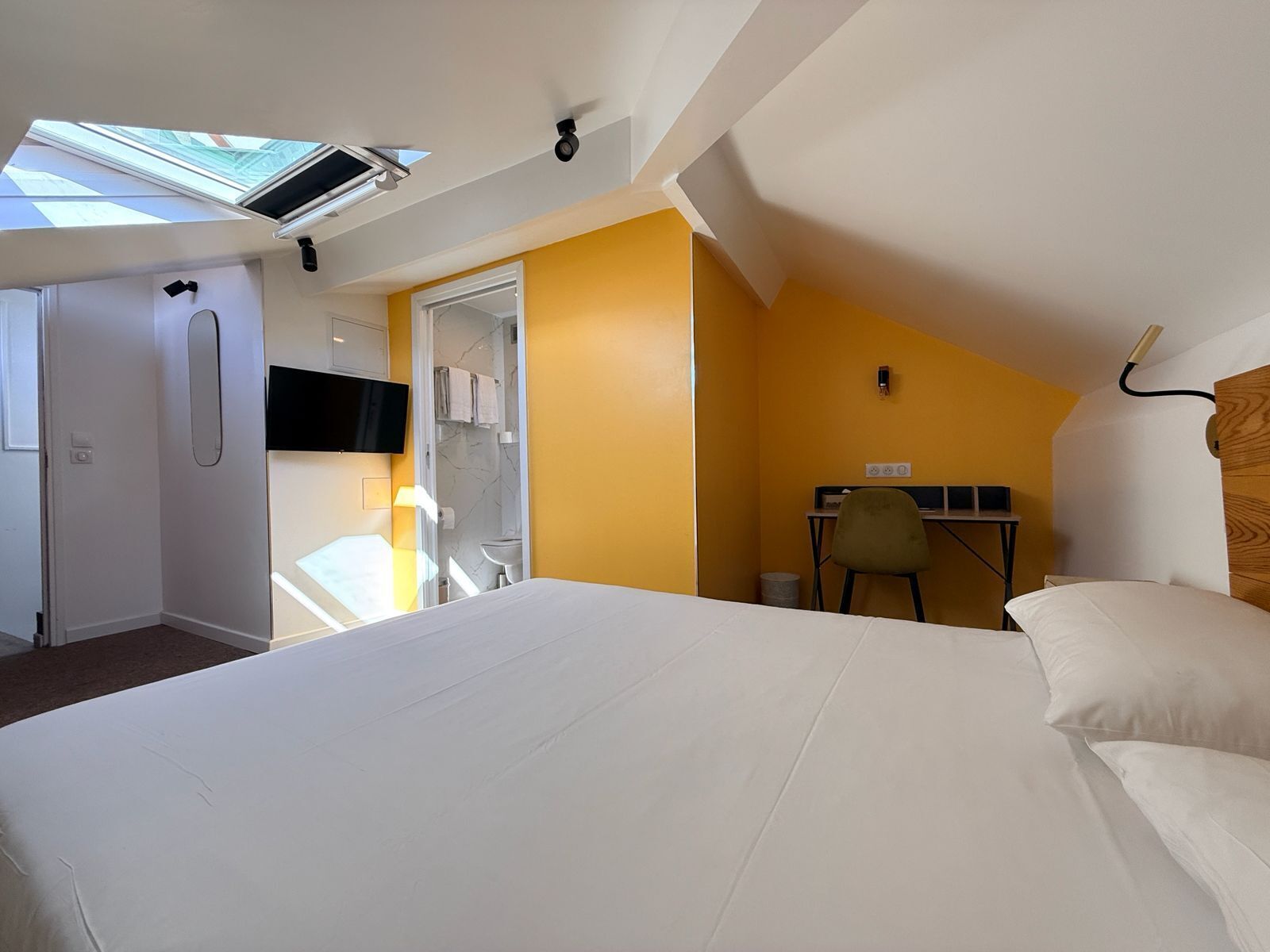 A bedroom with a bed, a desk, and a chair. There is a skylight above the bed and a door leading to a bathroom.
