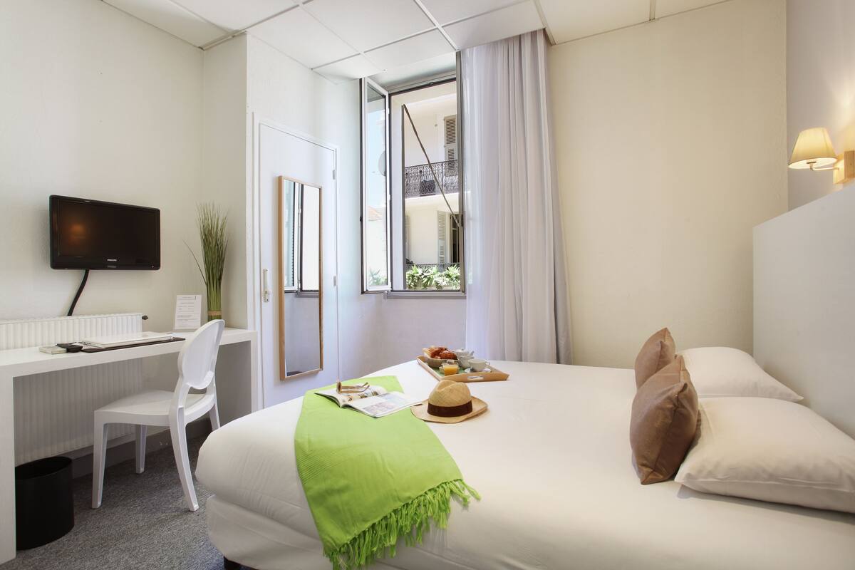 Ein Hotelzimmer mit einem Bett, einem Schreibtisch, einem Stuhl, einem Fernseher und einem Vorhangfenster.