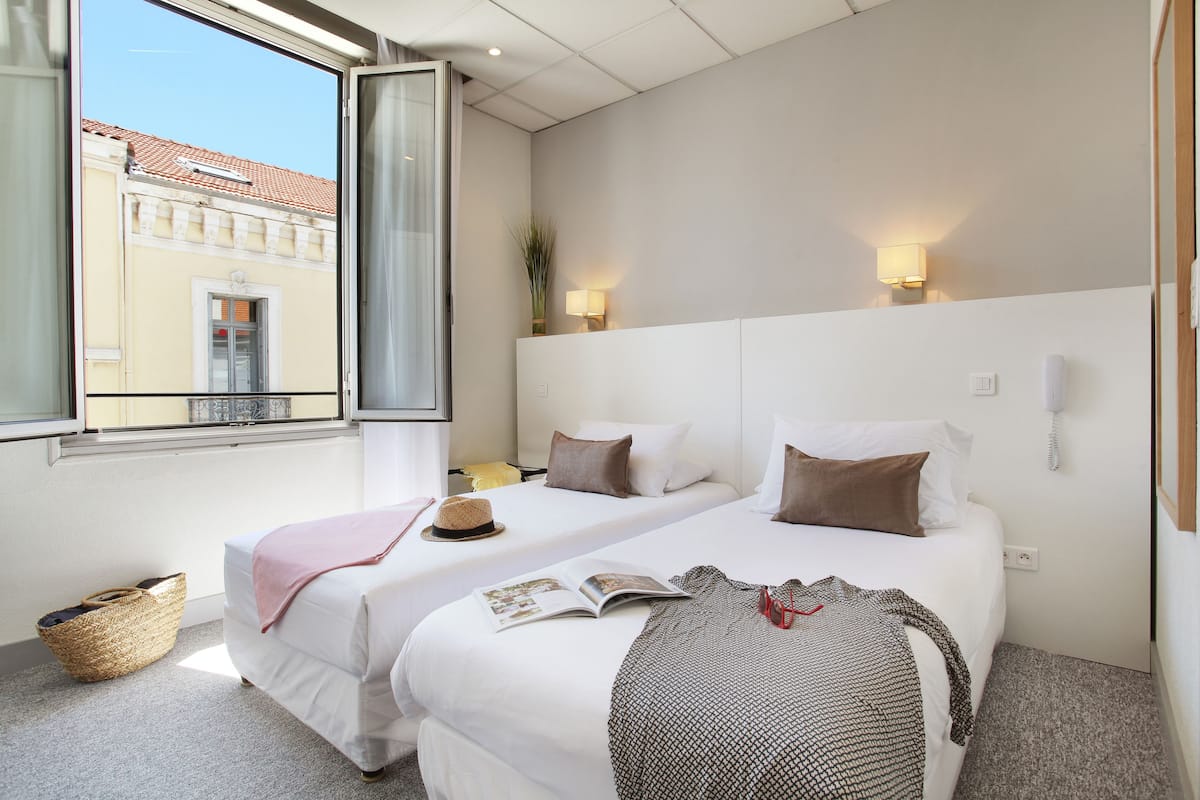 Ein Hotelzimmer mit zwei Betten, einem Fenster mit Blick auf Gebäude, einer Lampe und einem Telefon an der Wand.