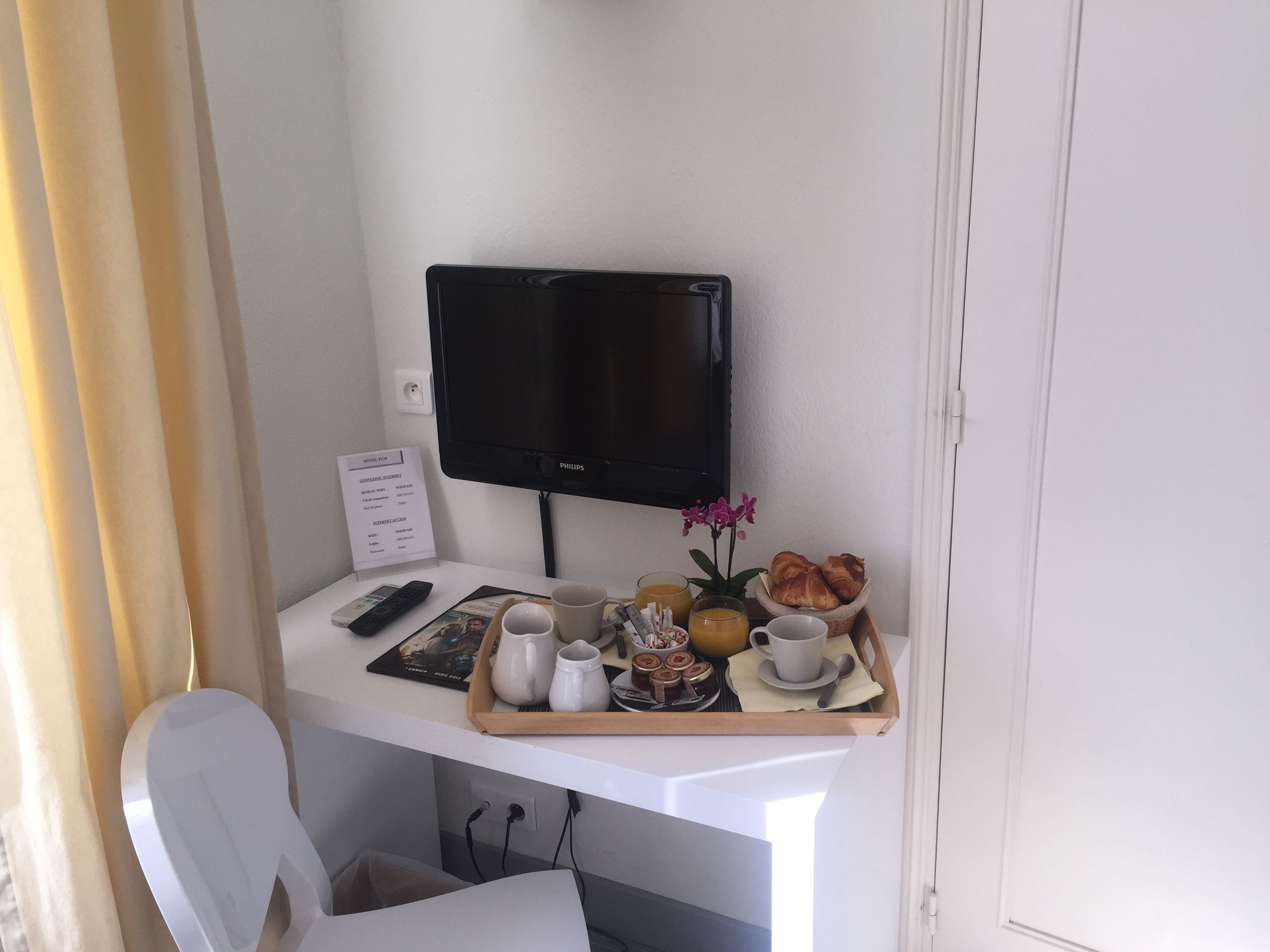 A hotel room with a white desk, a flat-screen TV, a tray with breakfast items, a remote control, and a menu sign.