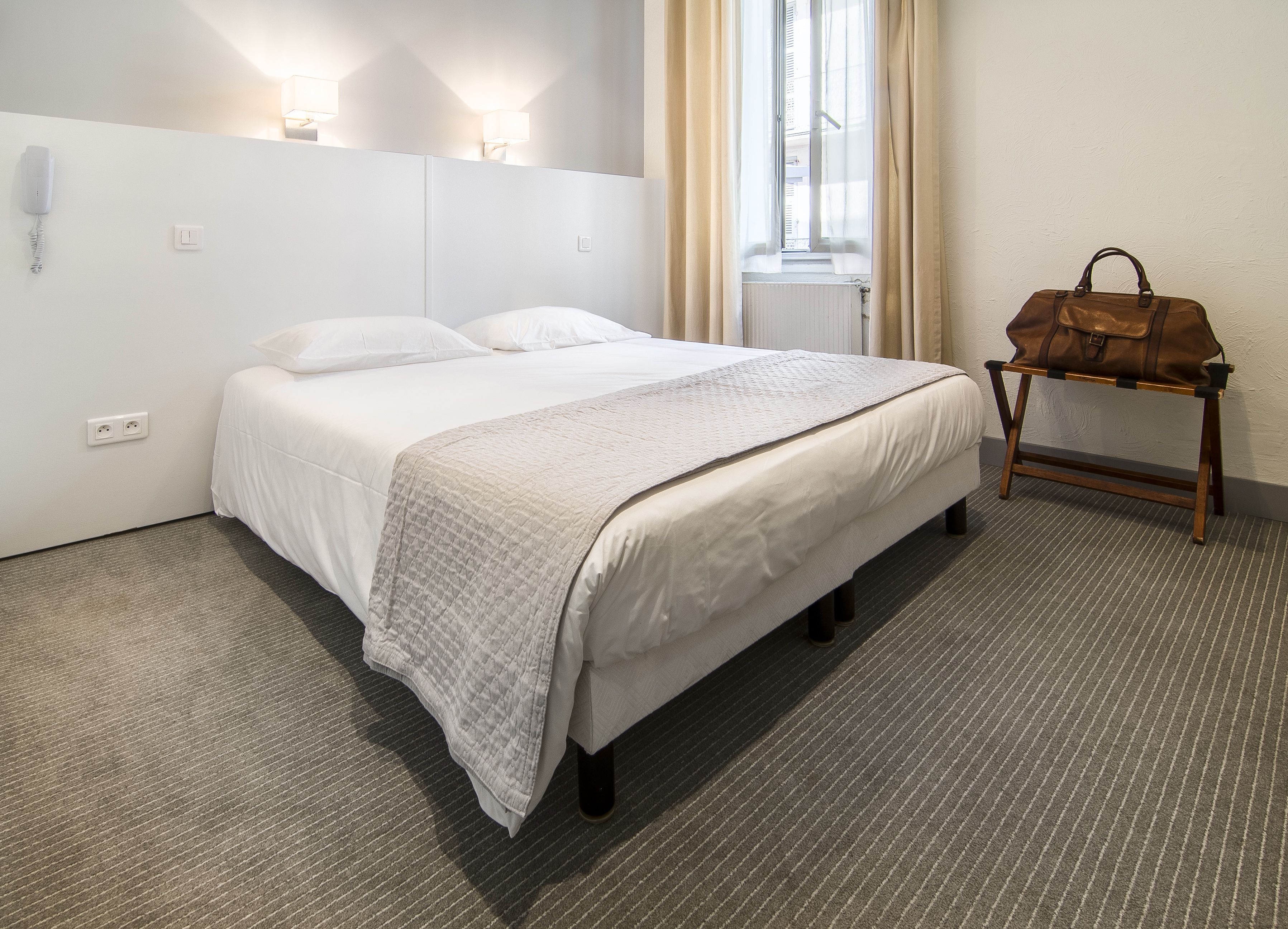 A hotel room with a bed, a bedside table, a lamp, and a window with curtains.